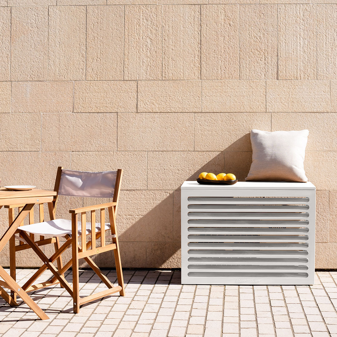 Cache clim blanc crème (RAL 9010) – taille S, installé avec soin – vue de face avec déco. Finition en aluminium haut de gamme, conçue pour habiller votre clim ou pompe à chaleur avec style. Un cache clim design pensé pour durer.