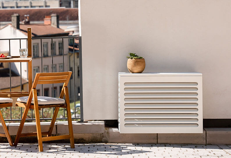 Cache clim taille XS Cabanaclim blanc installé sur un balcon de ville, avec mobilier en bois et jardinière décorative