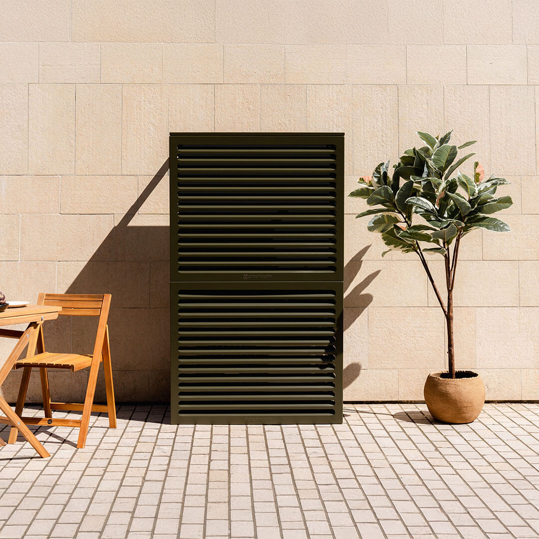 Double cache clim Cabanaclim vert olive dans un patio ensoleillé, devant un mur beige avec mobilier et plante