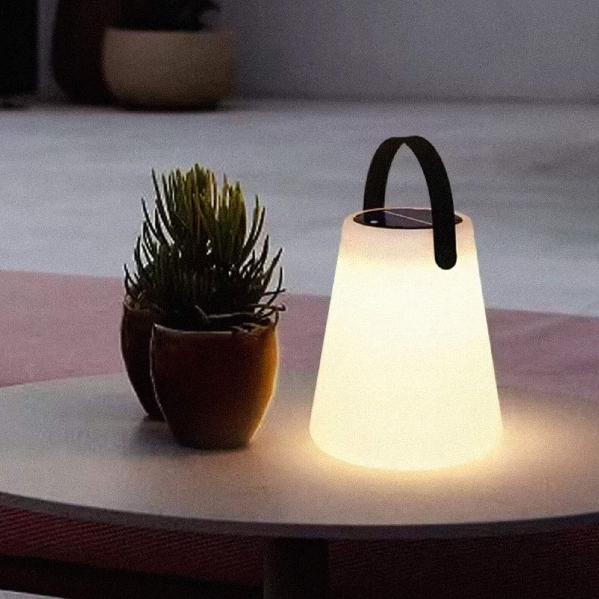Lampe solaire d’extérieur design avec poignée noire, posée sur une table ronde avec plante en pot, ambiance nocturne